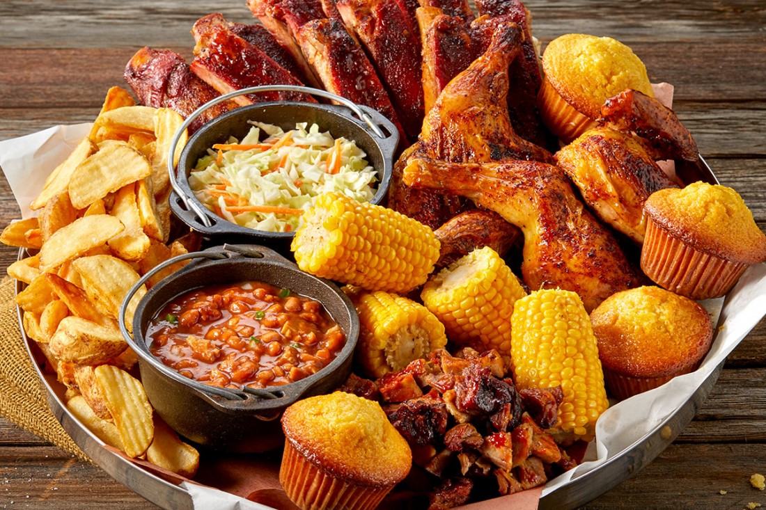 The Flying Pig Feast with chicken wings, ribs, cornbread muffins, celery sticks, pickles, and a bowl of mac and cheese piled high on a black platter