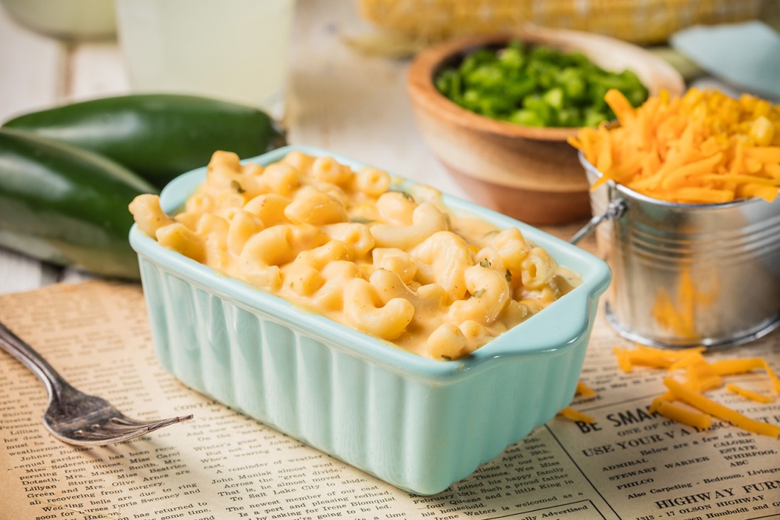 Dave's Cheesy Mac & Cheese, served in a small, light turquoise casserole dish. Behind it sits two whole jalapenos, a ramekin of chopped green onions and shredded cheese in a small pail