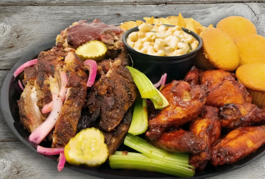 The Flying Pig Feast with chicken wings, ribs, cornbread muffins, celery sticks, pickles, and a bowl of mac and cheese piled high on a black platter