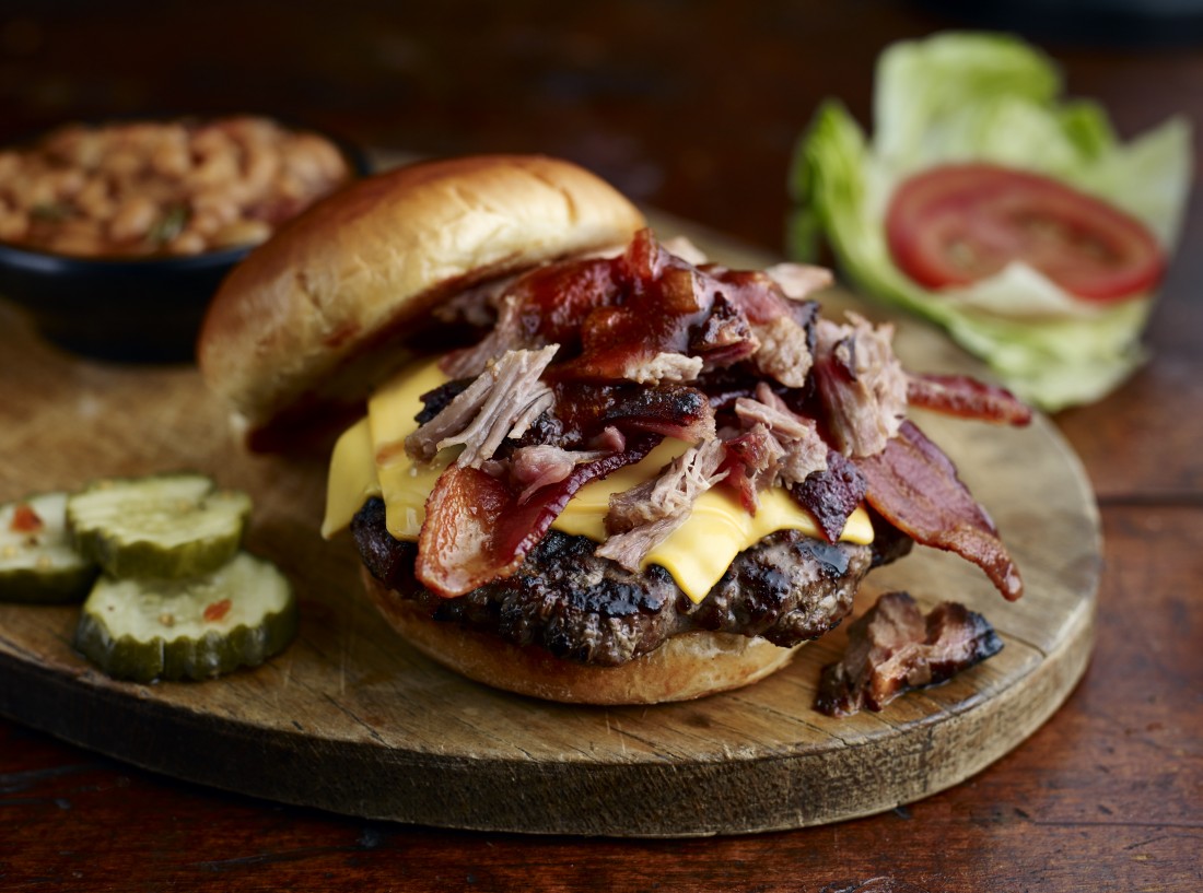 Famous Dave's BBQ Legendary Burger with chopped meat and a side of pickles, baked beans are in the background