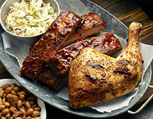 Famous Dave's BBQ ribs and roasted chicken on a grey platter, with bowls of coleslaw and baked beans on the side