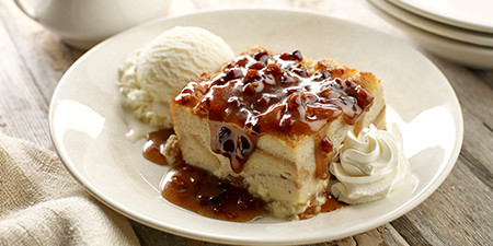 Famous Dave's Award-Winning Bread Pudding on a white plate, topped with a golden brown pecan praline sauce, with a scoop of vanilla ice cream on the side